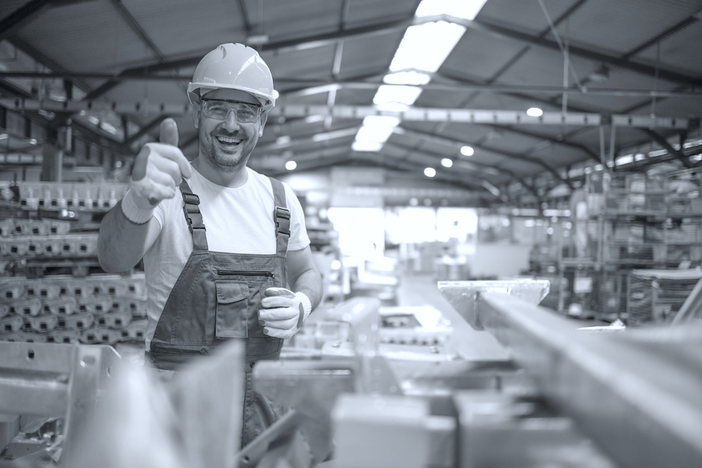 Portrait of factory worker in protective equipment holding thumbs up in production hall.